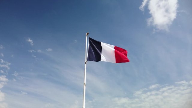 French flag flaps in the wind from a flagpole with blue sky behind it.
