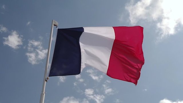 Slo-mo clip of French flag flapping in the wind in front of blue sky with scattered clouds.
