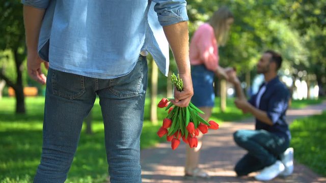 Lonely man with flowers watching male proposing marriage to his beloved girl