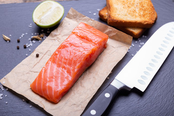 Salmon, avocado, lime and toasts laying with knife on slate background.