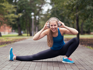 Blond smiling girl in the headphones doing stretching in the park outdoor, selective focus
