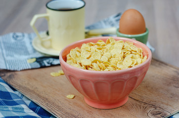 Corn flakes in a pink bowl, a mug with tea and boiled egg, on a wooden stand. copy space