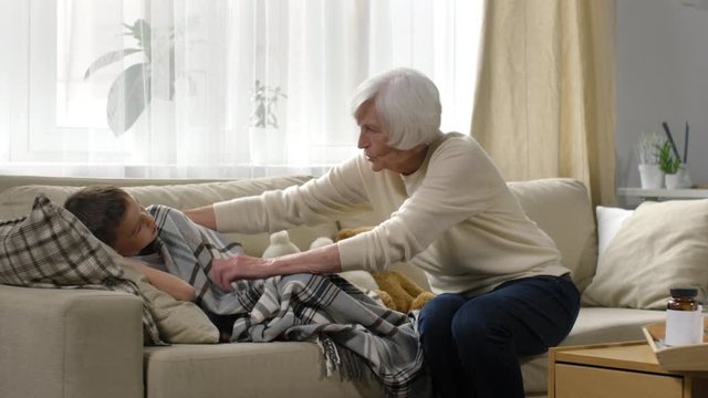Medium shot of caring grandmother with grey hair covering sick boy with warm blanket and putting her hand on his forehead to check temperature