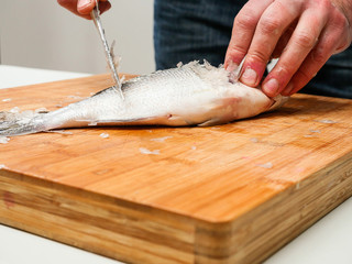 Descaling fresh sea bream on a wooden board with a sharp knife.
