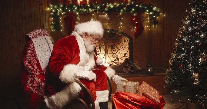 Authentic santa claus sitting in his rocker near fireplace, looking at camera and offering a christmas gift - christmas spirit, holidays and celebrations concept 4k footage
