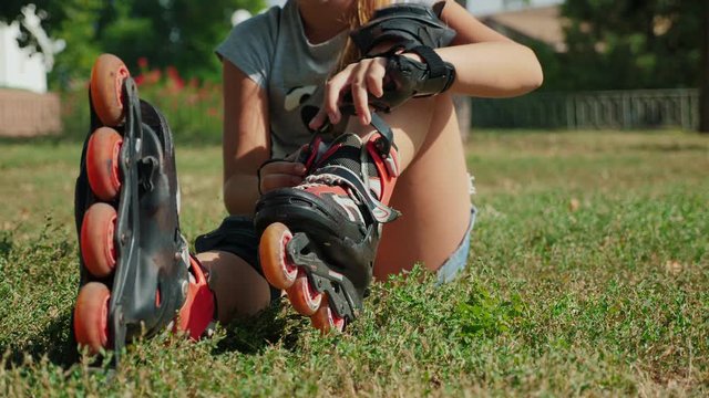 Young teenager girl sitting on the grass in a city park putting on her rollerblades - closing up the clasp. Close-up 4k video