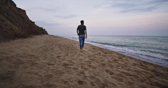 A charming man with a beard walks on the beach slowly and admiring the waves as they clash on the sand