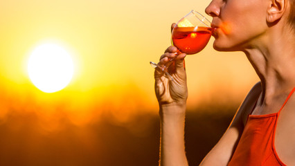 Happy brunette woman holding orange cocktail over sunset sky in orange colors