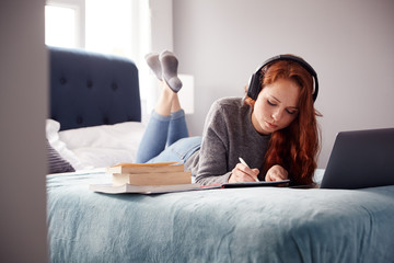 Female College Student Wearing Headphones Lies On Bed In Shared House With Mobile Phone Using Laptop - obrazy, fototapety, plakaty