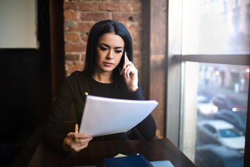 Serious woman financier exploring paper documents with new contract, sitting in restaurant after work day. Female skilled business owner reading summary during cellphone conversation