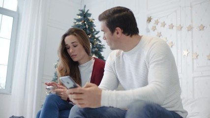 Serious couple sitting with mobile phones in luxury bedroom.