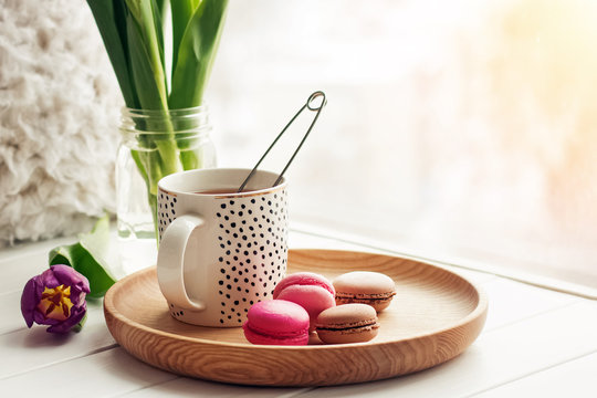 A cup of tea, sweet dessert macarons and purple tulips on a window sill.