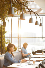Young businesswoman in casual clothing sitting at the table and typing on laptop computer with man sitting in the background in cafe