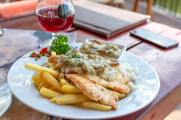 Grilled chicken fillet in white mushroom sauce with french fries on white plate. Glass of red wine in background