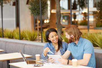 Young smiling businesswoman talking to the businessman and pointing at digital tablet in his hand during their meeting in cafe