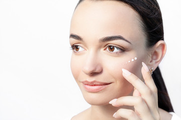 Beautiful model applying cosmetic drops of cream on her face on white background close-up