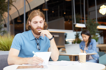 Serious bearded businessman in casual clothing sitting at the table and writing his future plans in notepad with woman working on laptop