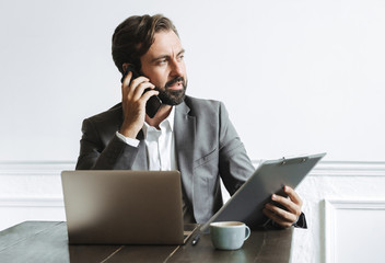 Image of thinking focused businessman holding clipboard and talking on cellphone while working in office