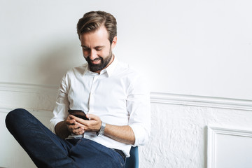 Image of pleased attractive businessman typing on cellphone and sitting on chair while working in office