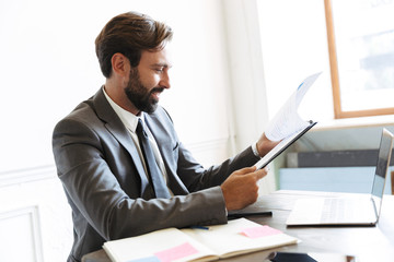 Image of smiling handsome businessman reading documents while working at laptop in office