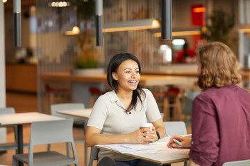 Happy young woman drinking coffee and talking to the young man while they sitting at the table and discussing working moments in cafe