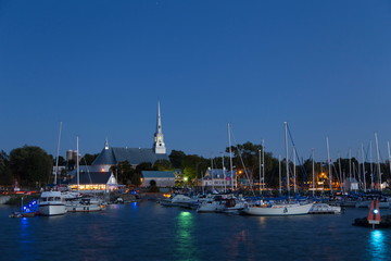 Fototapeta na wymiar Saint-Michel-de-Bellechasse marina seen during an early evening summer blue hour, with sailboats, historical 1873 church and waterside restaurant in the background, Quebec, Canada