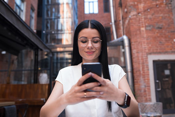 Stylish hipster girl with dark hair wearing in trendy spectacles online banking via mobile phone while relaxing in coffee shop. Female online shopping store via cell telephone