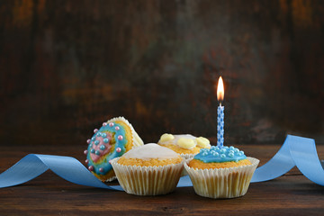 decorated muffins or cupcakes with a blue ribbon and a burning candle on a rustic wooden board against a dark brown background with copy space