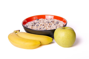 Sprouted wheat porridge with raisins and dried apricots in red and black bowl with green apple and yellow bananas near it. Isolated on white background. Idea for healthy diet breakfast.