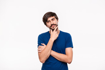 Thoughtful handsome brunet man in a blue shirt holding arms folded over his chest and keeping one hand close to face standing isolated over white background. Emotion of idea and thinking.