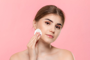 Beautiful model applyes cosmetic product to skin with white cotton sponge. Model looks at the camera isolated over pink background. Concept of beauty and health treatment.