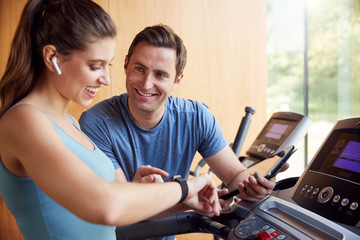 Woman In Gym With Personal Trainer Analysing Performance Using Smart Watch And Digital Tablet