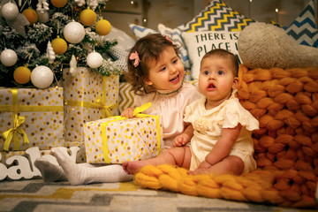 Two sisters girls sitting on an orange warm plaid in dresses under a Christmas tree with gifts, joyful childhood