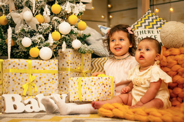 Fototapeta premium Two sisters girls sitting on an orange warm plaid in dresses under a Christmas tree with gifts, joyful childhood