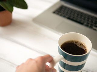 Woman's hand touches the large mug of coffee on the white wooden desk. Notebook and green plant on background.