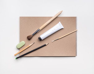 Composition with open craft paper sketchbook, pencils, brushes and tube of paint on white background
