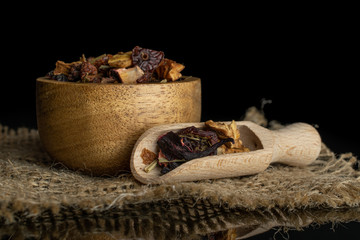 Lot of pieces of lingonberry strawberry fruit tea in wooden bowl with wooden scoop on jute cloth isolated on black glass