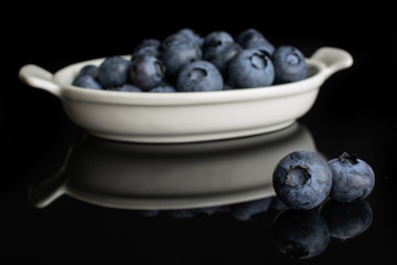 Lot of whole fresh blue bilberry in white oval ceramic bowl isolated on black glass