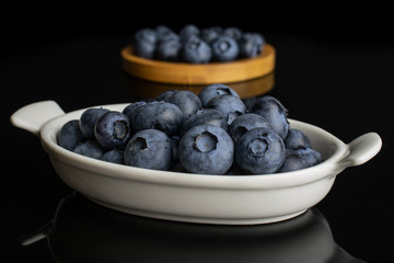 Lot of whole fresh blue bilberry on round bamboo coaster in white oval ceramic bowl isolated on black glass