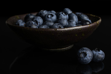 Lot of whole fresh blue bilberry in dark ceramic bowl isolated on black glass