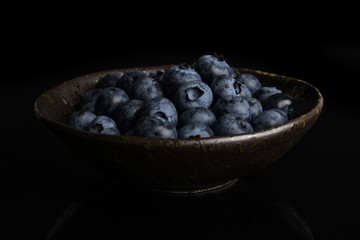 Lot of whole fresh blue bilberry in dark ceramic bowl isolated on black glass