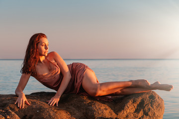 Sexy tanned woman posing reclining on coastal rocks. The sea in the background. Sunset light