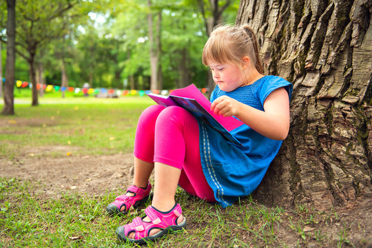 A portrait of trisomie 21 child girl outside having fun on a park reading book