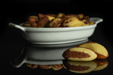 Lot of whole raw brown brazil nut in white oval ceramic bowl isolated on black glass