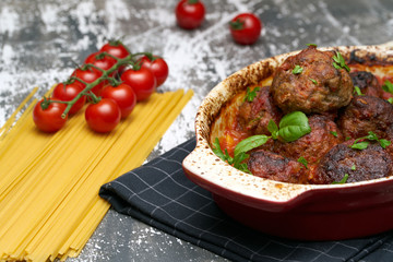 meatballs with tomato sauce and decorated with basil leaf, served in white and red pan on grey background. next to the pan near fresh tomatoes, spaghetti. Closeup.