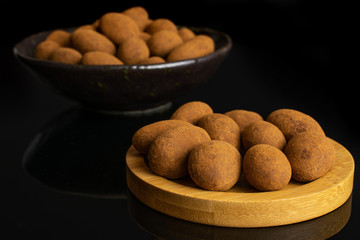 Lot of whole sweet brown chocolate cinnamon almond on round bamboo coaster in dark ceramic bowl isolated on black glass