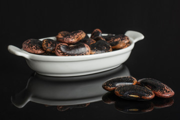 Lot of whole stained purple bean in white oval ceramic bowl isolated on black glass