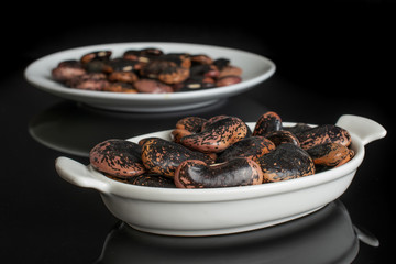 Lot of whole stained purple bean in white oval ceramic bowl on white ceramic plate isolated on black glass
