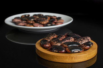 Lot of whole stained purple bean on round bamboo coaster on white ceramic plate isolated on black glass