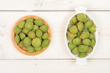 Lot of whole spicy green wasabi peanut in white oval ceramic bowl on bamboo coaster flatlay on white wood